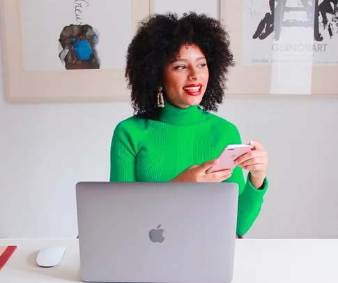 Person in a bright green sweater smiling, holding a phone, sitting at a desk with a laptop.