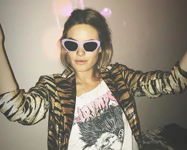 Person wearing pink sunglasses, zebra-pattern jacket, and graphic t-shirt, arms raised in a playful pose.