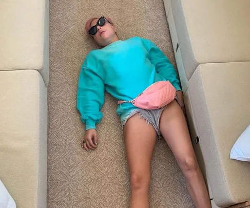 Person with pink hair in a teal sweater and denim shorts, lying on the floor between sofas, wearing sunglasses.