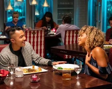 A couple engaged in conversation at a restaurant, seated at a table with plates and drinks, in a cozy, ambient setting.