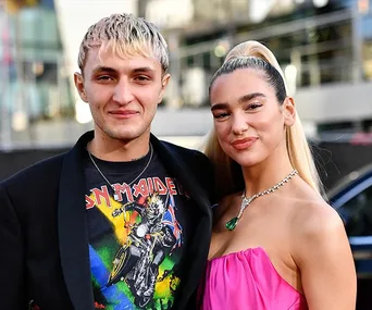 A smiling couple poses for a photo; the woman is in a pink dress, and the man wears a band t-shirt and jacket.