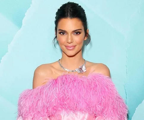 Person in a stylish pink outfit against a turquoise background, wearing a diamond necklace, smiling at the camera.