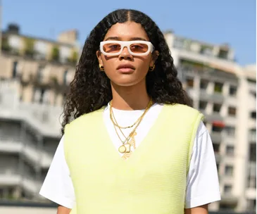 Person wearing a yellow sweater vest over a white t-shirt, accessorized with white sunglasses and gold jewelry.