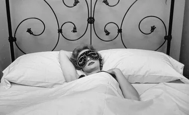 Person lounging in bed with an ornate headboard, wearing a sleep mask and relaxing under a blanket.