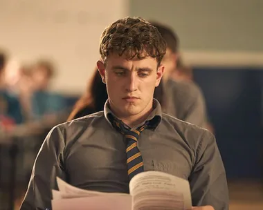 Man in a gray shirt and striped tie, intensely reading papers, set in a classroom environment.