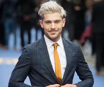 A man with blond hair in a plaid suit and orange tie, smiling at a formal event.