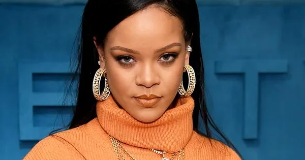 Rihanna in orange turtleneck sweater with large hoop earrings, posing against a blue backdrop, promoting Fenty Beauty.