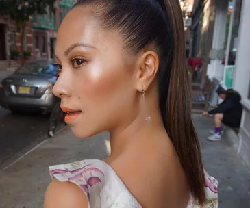 Woman with highlighted cheeks, ponytail hairstyle, wearing earring, glances over shoulder on a city street.