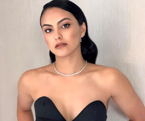 Woman with long dark hair, winged eyeliner, and a strapless black top against a light-colored wall.