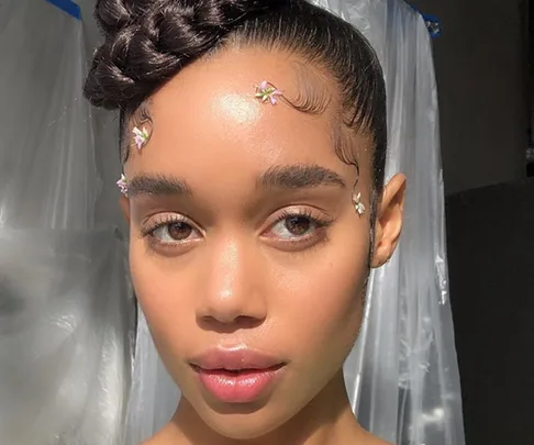 A person with styled baby hairs adorned with small flowers, wearing a braided updo, in front of a sheer backdrop.