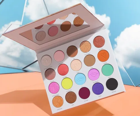 Open eyeshadow palette with 18 vibrant colors, placed on an orange surface, with blue sky and clouds in the background.