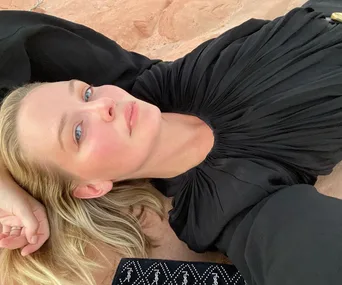 Person in black outfit lying on the ground, looking relaxed; blonde hair, soft lighting.