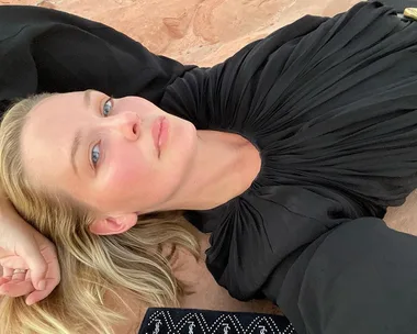 Person in black outfit lying on the ground, looking relaxed; blonde hair, soft lighting.