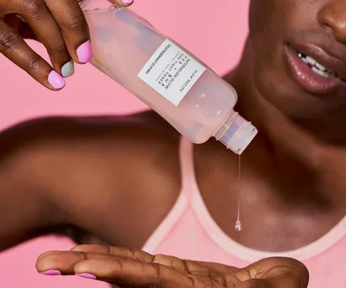 A person pours a skincare toner into their hand against a pink background.