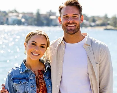 A smiling couple posing in sunlight by a waterfront, surrounded by blurred cityscape and water reflections.