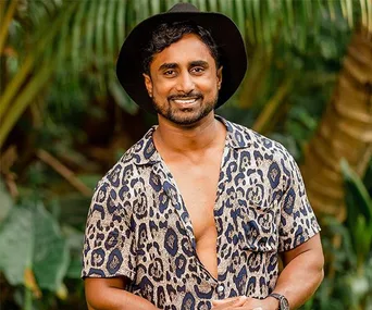 Man smiling outdoors in a patterned shirt, wearing a black hat, with greenery in the background.