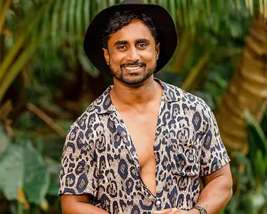 Man smiling outdoors in a patterned shirt, wearing a black hat, with greenery in the background.