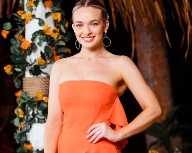 Woman in orange dress smiles, standing next to floral decor on "Bachelor in Paradise Australia" set.