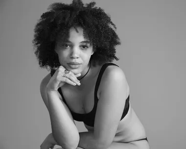 A person with curly hair in a contemplative pose, wearing a black bra top against a plain backdrop.