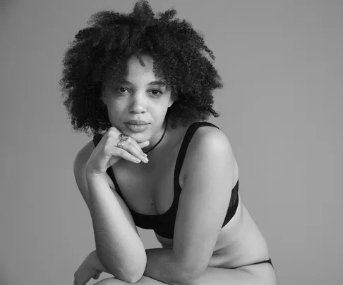 A person with curly hair in a contemplative pose, wearing a black bra top against a plain backdrop.