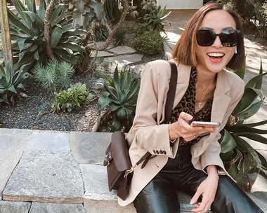 A woman in sunglasses smiles while sitting outside, holding a phone, with plants in the background.