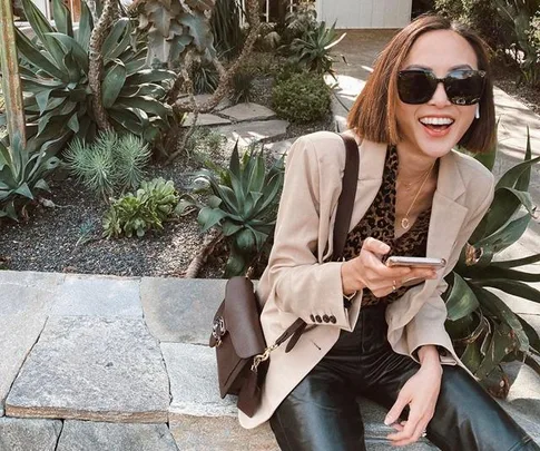 A woman in sunglasses smiles while sitting outside, holding a phone, with plants in the background.