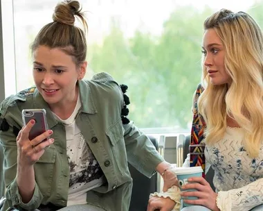 Two women are sitting; one looks surprised at her phone, the other holds a drink, indoors with a window view.