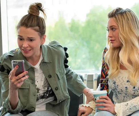 Two women are sitting; one looks surprised at her phone, the other holds a drink, indoors with a window view.