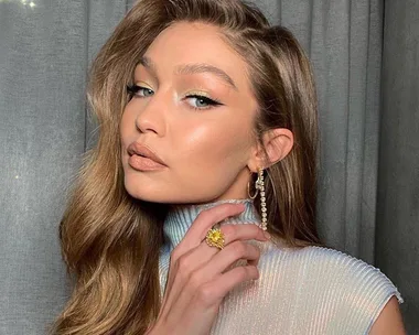 Gigi Hadid with wavy hair, wearing a metallic top, large gold earrings, and a statement ring.