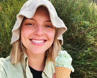 A person with a beige hat smiles while holding a mint ice cream cone, surrounded by green foliage.