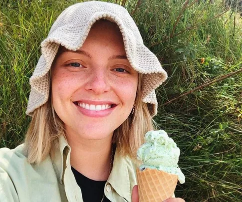 A person with a beige hat smiles while holding a mint ice cream cone, surrounded by green foliage.