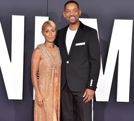 A couple posing on a red carpet; the woman in a golden dress and the man in a black suit, smiling at the camera.