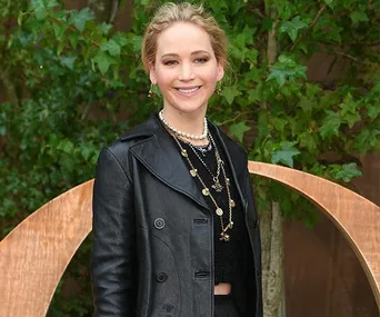 A woman in a black leather jacket with layered necklaces stands outdoors, smiling in front of greenery.