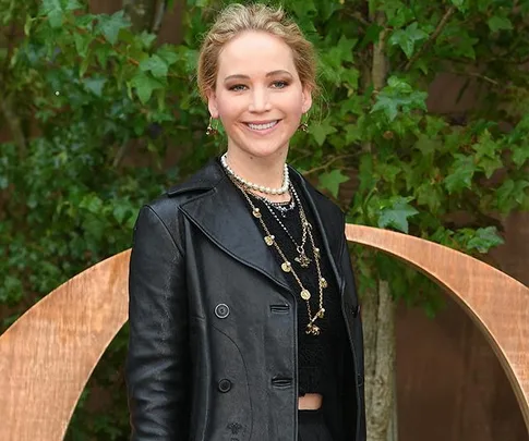 A woman in a black leather jacket with layered necklaces stands outdoors, smiling in front of greenery.