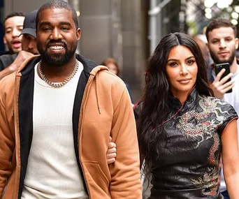 Two people walking outdoors, one in a light jacket and the other in a detailed black top, surrounded by a crowd.