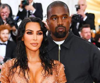 A woman in a sparkling dress and a man in a dark jacket stand together at a formal event, surrounded by photographers.