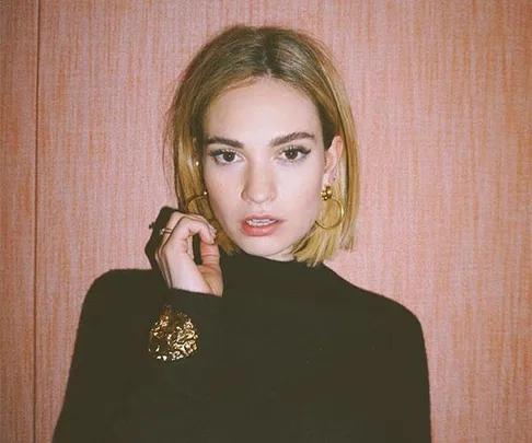 A person with short hair, wearing a black top and gold accessories, poses in front of a peach background.