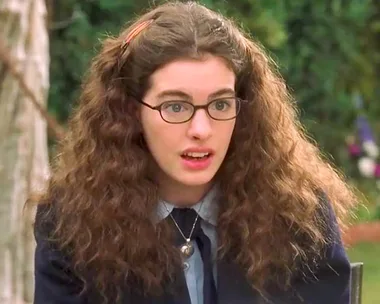 A young woman with frizzy hair and glasses looks surprised; she's in a garden setting.