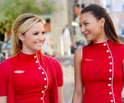 Two women wearing matching red uniforms with name tags "Dani" and "Santana," smiling at each other.