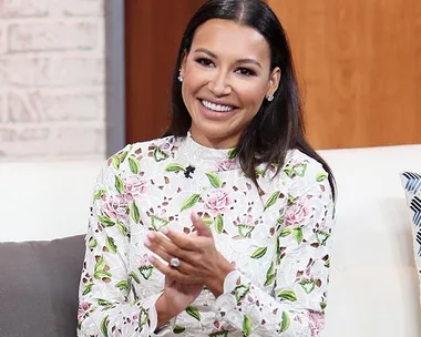 Smiling woman with long hair in floral top sitting, clapping hands, on a talk show set.