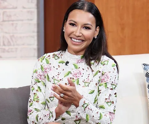 Smiling woman with long hair in floral top sitting, clapping hands, on a talk show set.