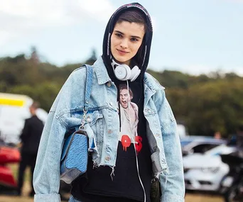 Person wearing a denim jacket, hoodie, and headphones, holding a blue bag outdoors.