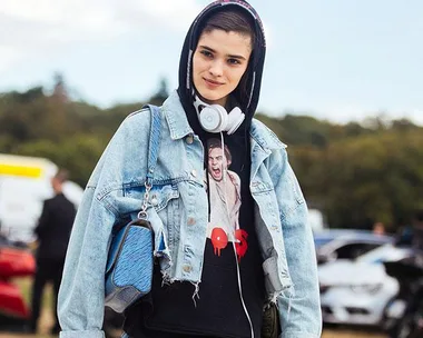 Person wearing a denim jacket, hoodie, and headphones, holding a blue bag outdoors.