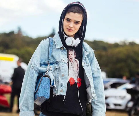 Person wearing a denim jacket, hoodie, and headphones, holding a blue bag outdoors.