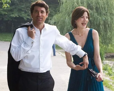 A man in a white shirt and woman in a blue dress laugh while walking in a park, with trees in the background.