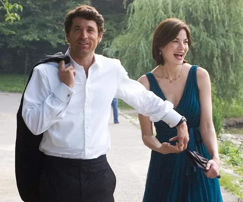 A man in a white shirt and woman in a blue dress laugh while walking in a park, with trees in the background.