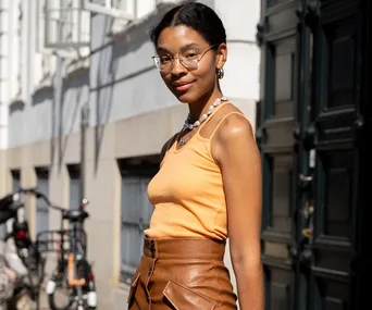 Woman in an orange tank top and brown leather skirt poses on a sunny street during Copenhagen Fashion Week.