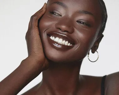 A woman with hoop earrings smiles, touching her face, showcasing smooth dark skin and glossy lips.