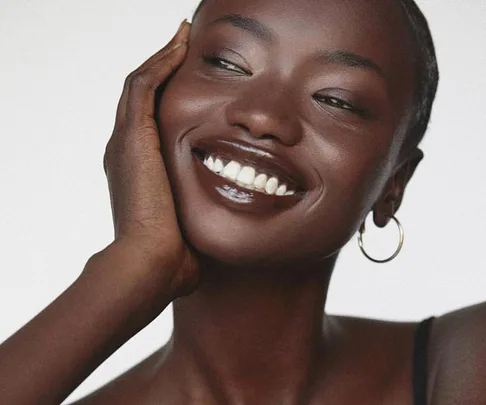 A woman with hoop earrings smiles, touching her face, showcasing smooth dark skin and glossy lips.