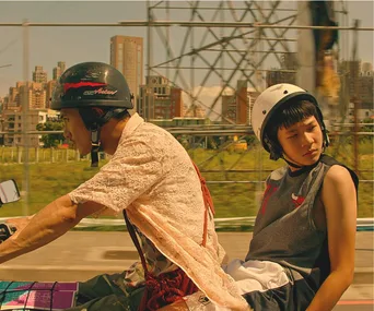 Two people on a motorcycle, wearing helmets, with an urban cityscape in the background.